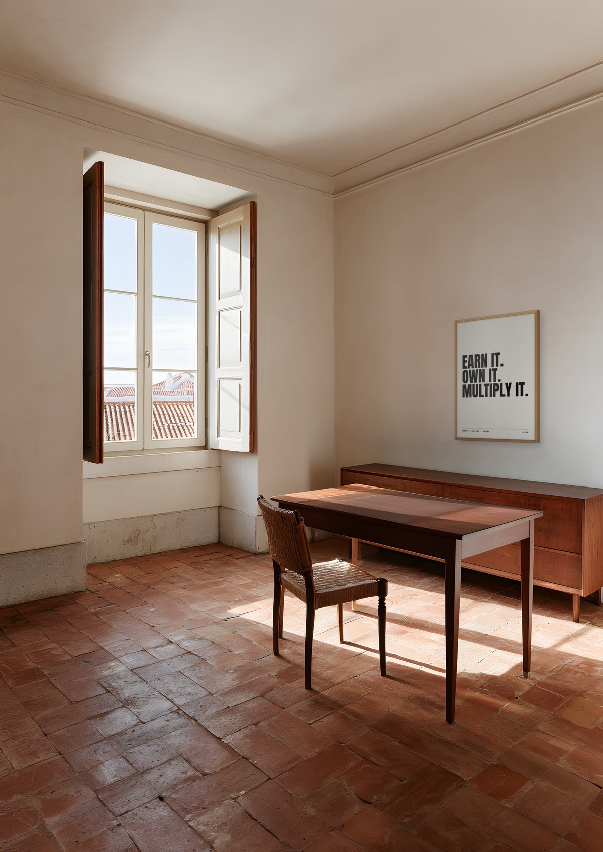 Earn It. Own It. Multiply It. in a natural wood frame, shown in a Lisbon apartment study